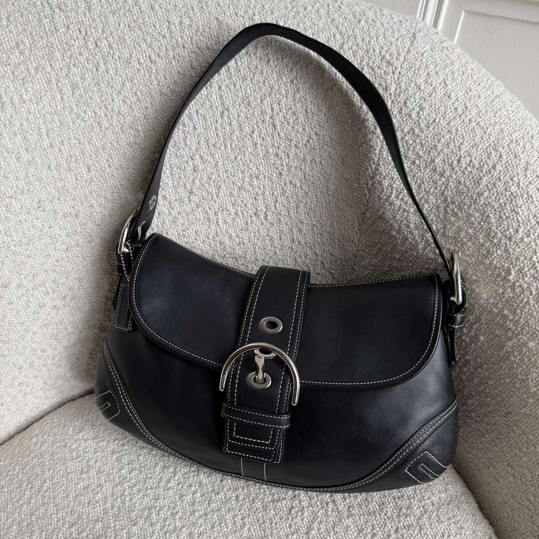 A black leather Coach Soho shoulder bag with a buckle design and a signature charm, placed on a textured surface.